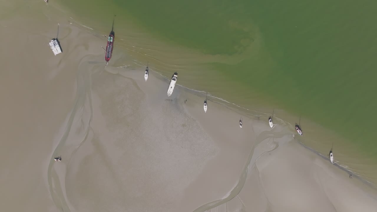 Vertical overhead of a motor yacht and small boats on the sand edge beside green water; high‑contrast survey of intertidal textures. Wadden Sea