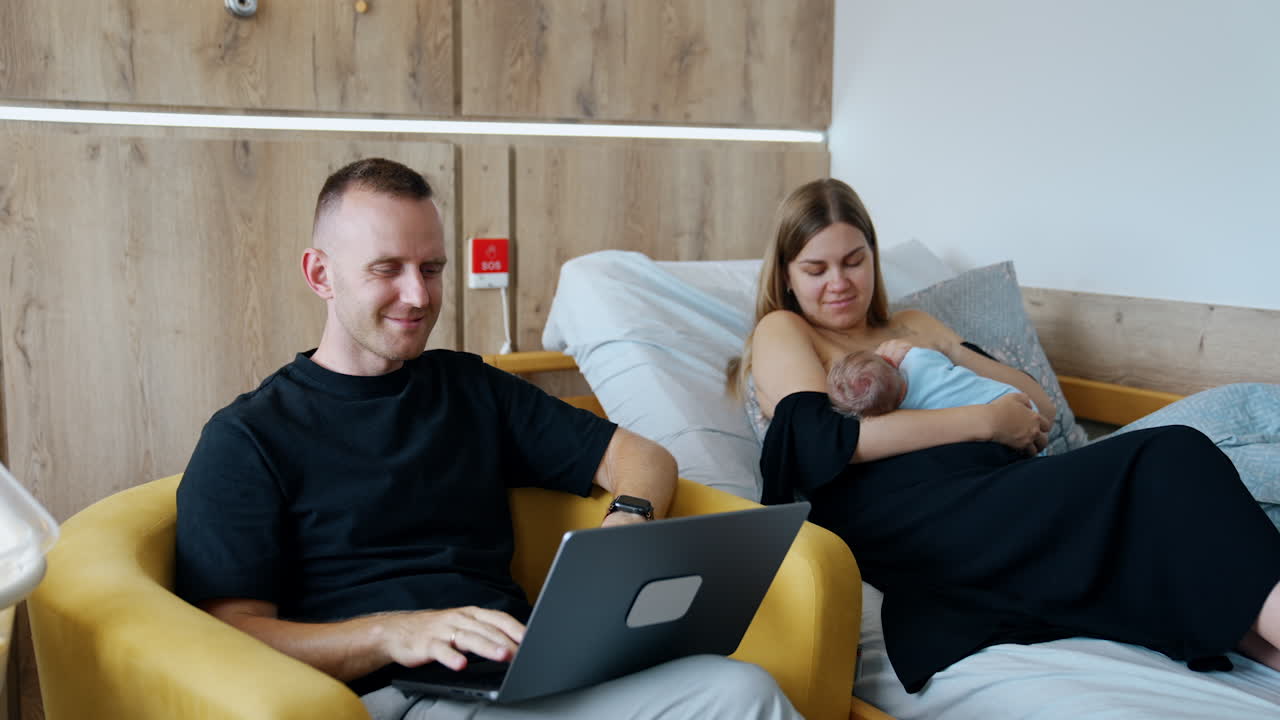 Modern family couple with a newborn in the hospital after childbirth. First time mom feeding a baby and dad listens to music on laptop.
