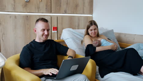 modern family couple with a newborn in the hospital after childbirth. first time mom feeding a baby and dad listens to music on laptop.