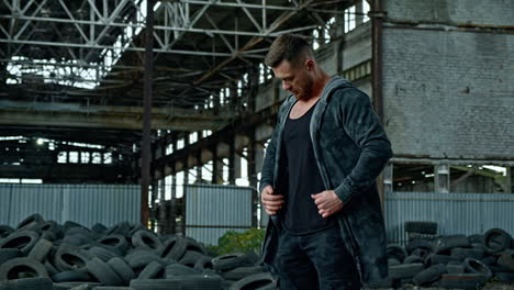 bearded man in military clothing stands in abandoned place. young man on the background of ruined plant with many old tires around.