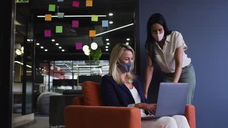 Diverse-businesswomen-wearing-face-masks-using-laptop-talking-in-office