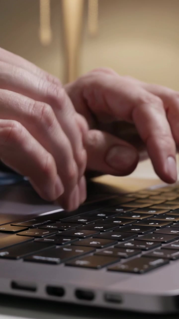 Vertical video: Typing with poised fingers on laptop keyboard at desk for work, with lamp