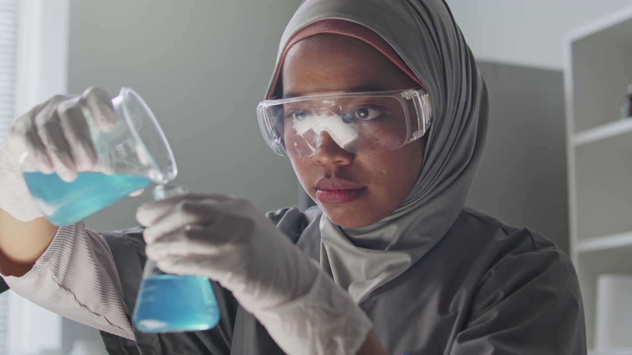 Ilmuwan Wanita Berhijab Menuangkan Cairan Biru dari Gelas Kimia ke Labu