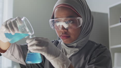 female scientist in hijab pouring blue liquid from beaker into flask