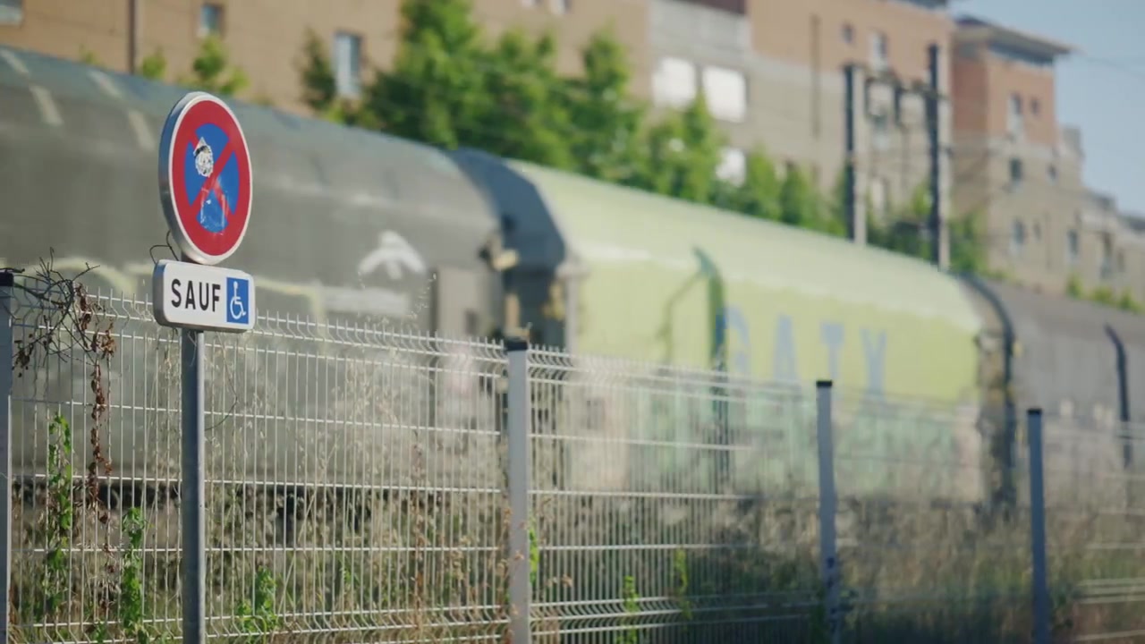 Close up of a roadside no-parking sign beside a passing train, with urban buildings and railway tracks blurred in the background
