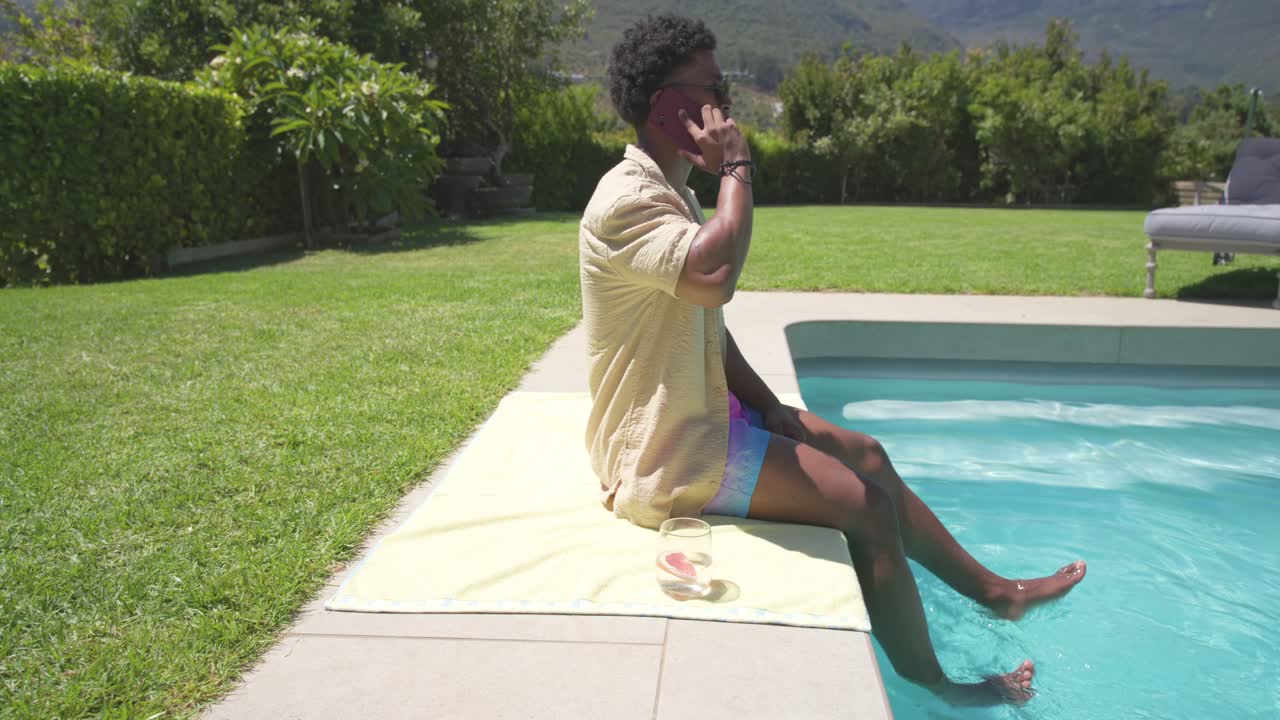 Smartphone klingelt, afroamerikanischer Mann nimmt am Pool auf Handtuch liegend ab, taucht Füße ins Wasser, Glas in der Hand, mit Kopierraum