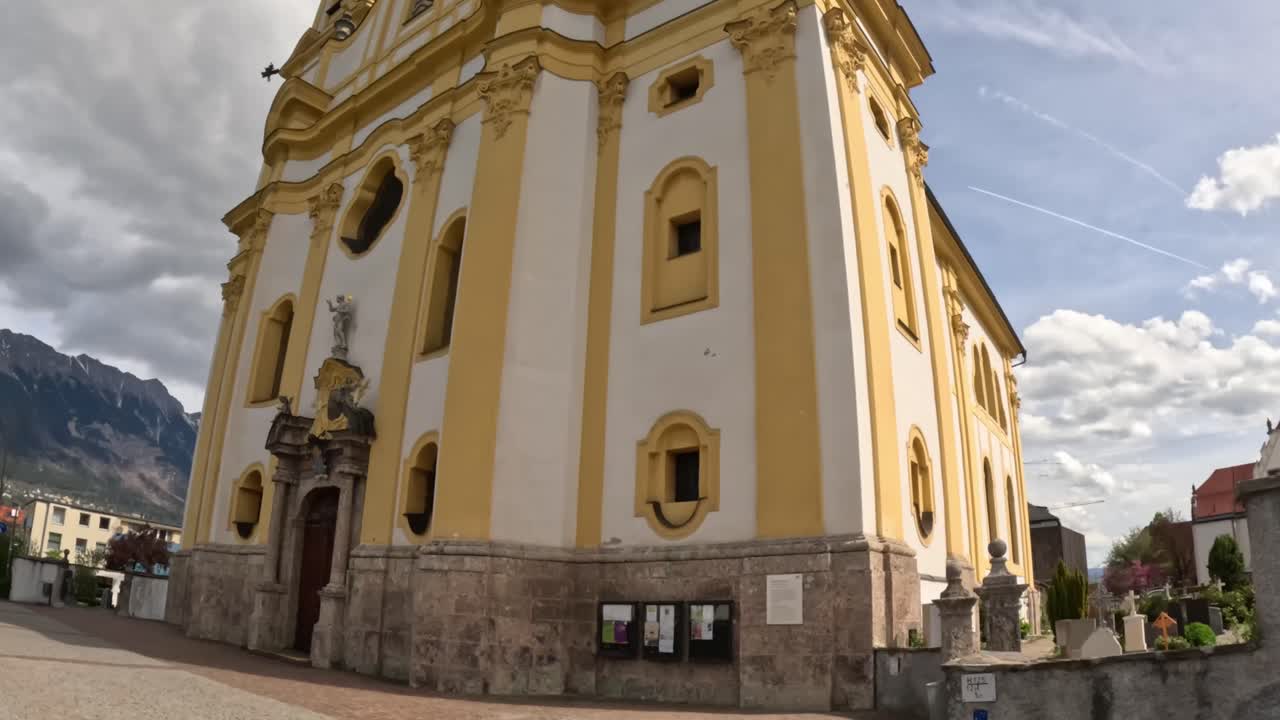 Wide angle view of the yellow and white baroque Pfarrkirche und Basilika Mariae Empfängnis in Innsbruck, Austria, showcasing its elegant architecture, stone base, and alpine backdrop, tilt up camera