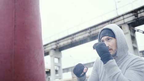 close-up view of caucasian man in grey hoodie hitting a punching bag outdoors an abandoned factory on a cloudy morning