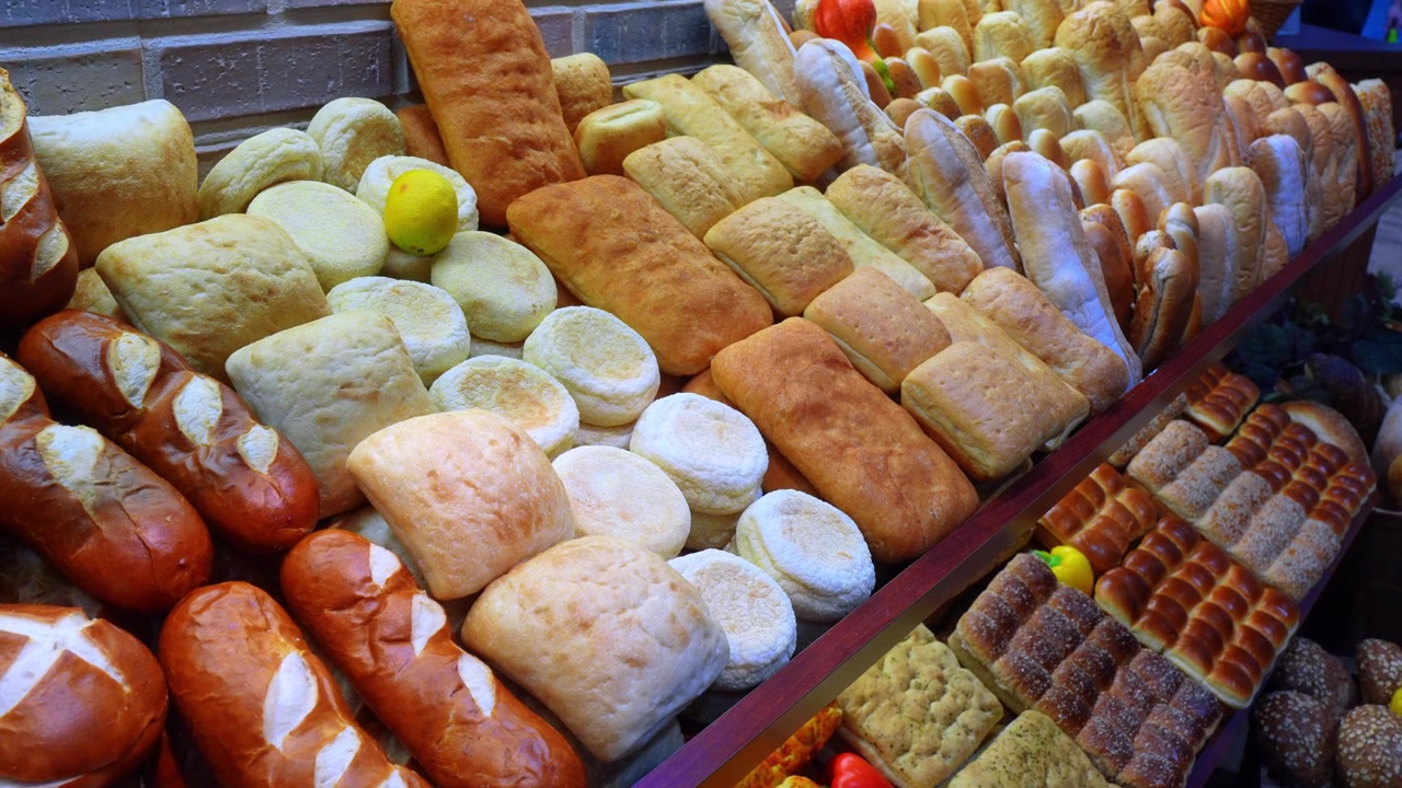 Bir pazar tezgahında parlak ışıklandırılmış geleneksel ekmek seçkisi. Simit rulolarını, yassı ekmekleri ve beyaz çörekler arasında tek bir sarı limonu sergileyen yakın çekim yukarıdan görünüm.