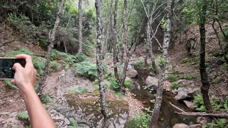 Chica-Morena-Con-Gafas-Tomando-Una-Fotografía-Con-Su-Teléfono-Celular-En-Un-Sendero-Con-Un-Río-En-Un-Día-Nublado