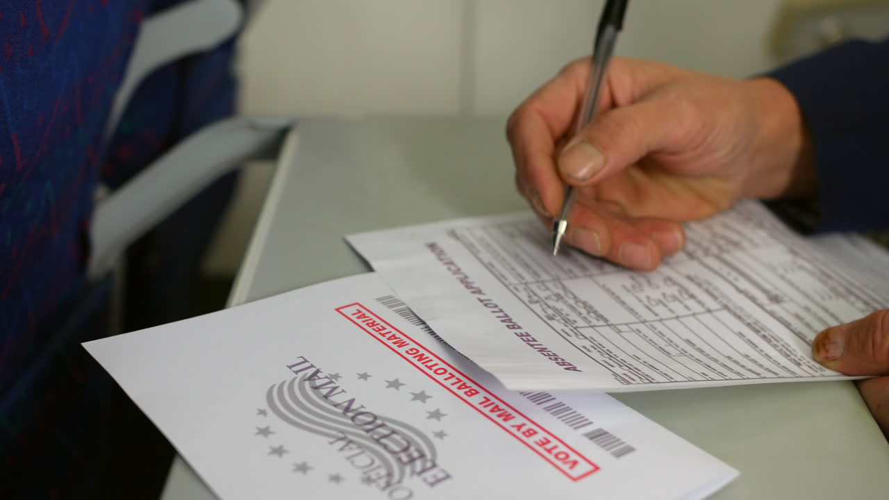 Filling out an absentee ballot application form with a pen. The close-up shot emphasizes the details of the form and the "Official Election Material" envelope, highlighting the voting process
