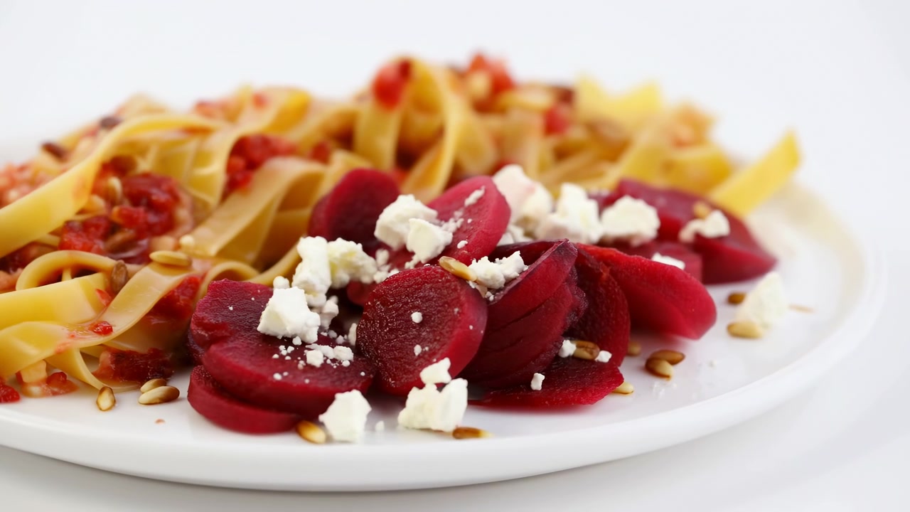 Tagliatelle con fette di barbabietola, feta sbriciolata, salsa di pomodoro e semi tostati, su piatto in ceramica bianca luminoso.