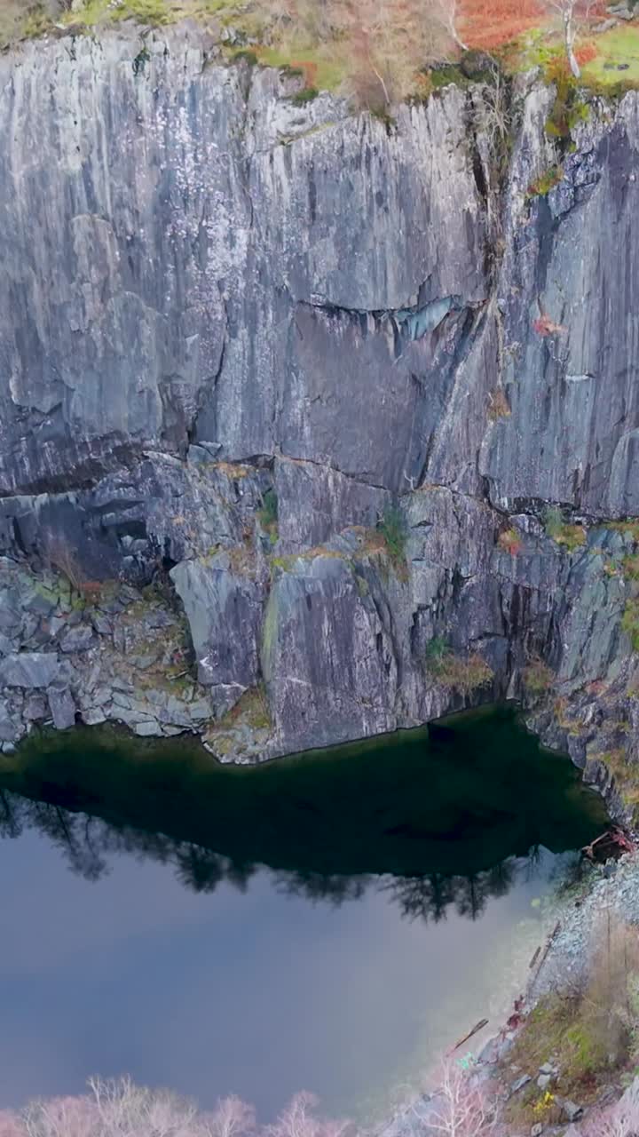 Discesa verticale di un drone verso un lago di cava, spostando la messa a fuoco dalle cime degli alberi e dai colori invernali in alto alle ripide pareti di ardesia e all'acqua ferma sottostante in una drammatica rivelazione verso il basso