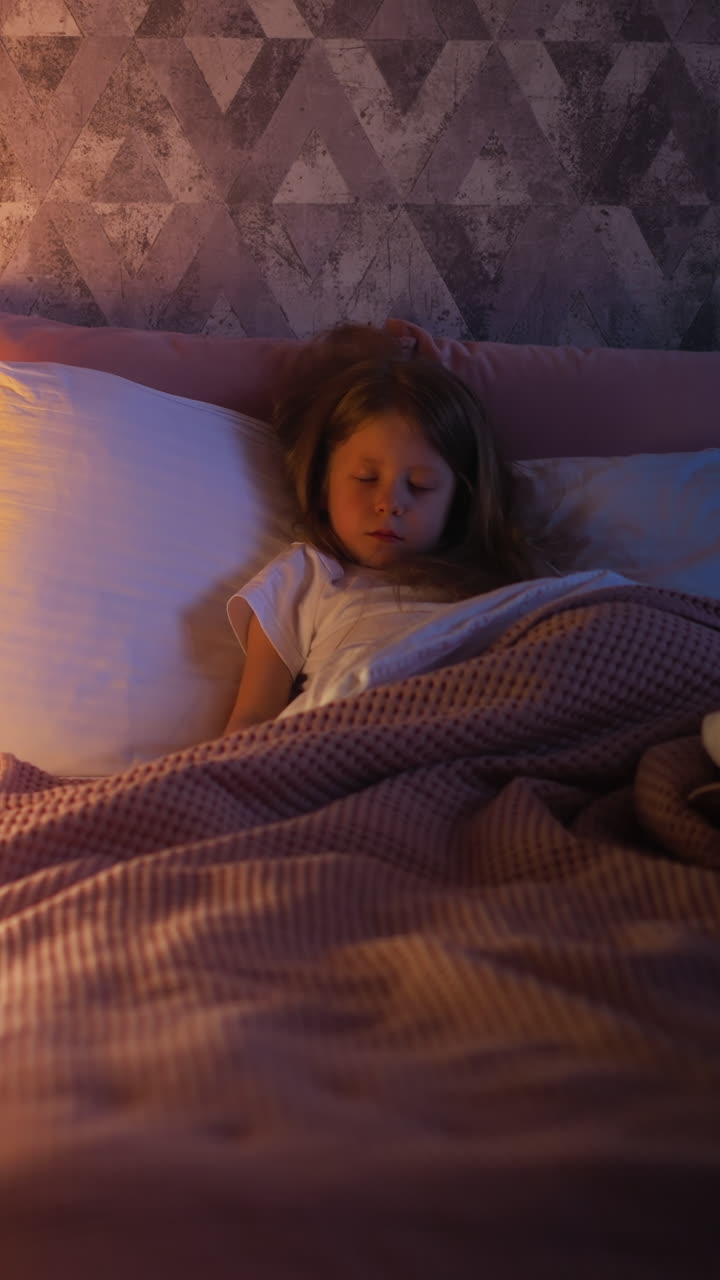 Adorable little girl sleeps tight lying on comfortable bed with warm plaid by illuminated children wigwam in bedroom with dark corner at home upper view slow motion