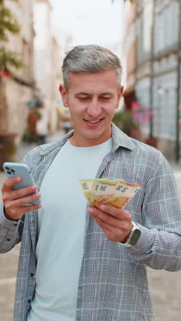 happy rich man counting holding money euro cash using smartphone calculator app on city street