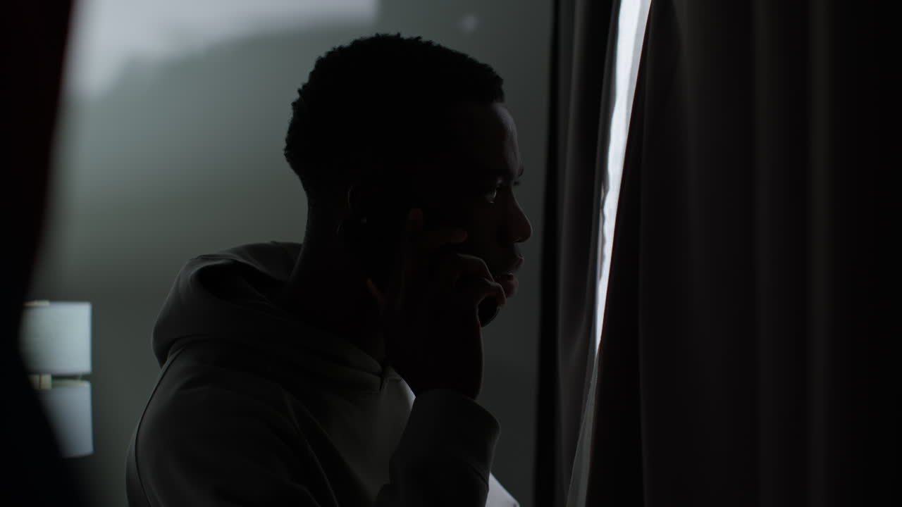 Young Man With Mental Health Issues Suffering With Paranoia Or Agoraphobia At Home Talking On Mobile Phone Looking Out Of Window 2