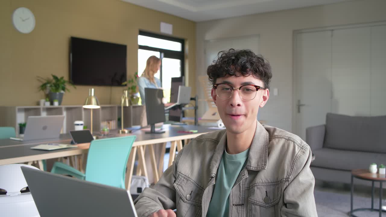 Male coworker typing at laptop on shared desk in office pausing smiling laughing as camera zooming