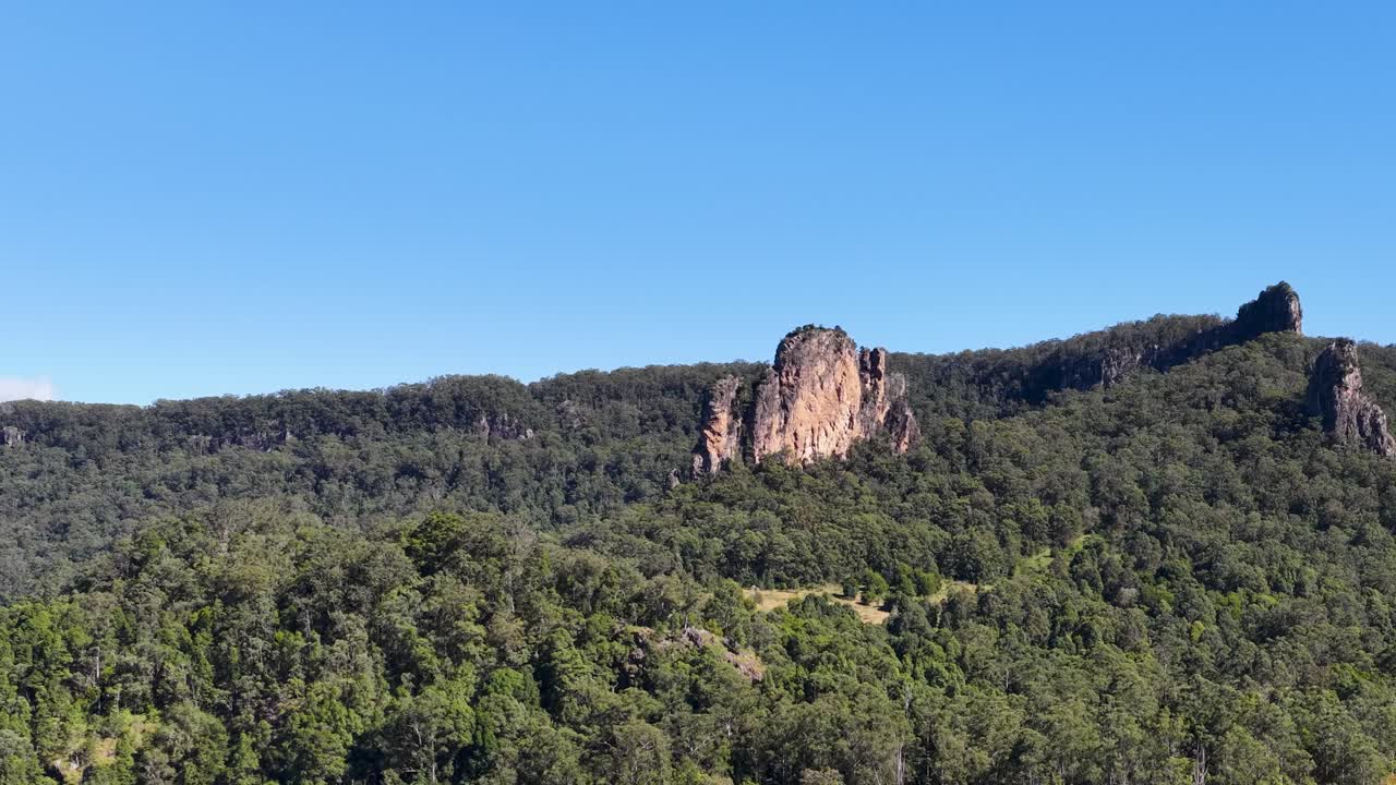 Panoramic view of Nimbin Rocks under clear blue skies. Lush greenery surrounds the iconic rock formations in bright daylight