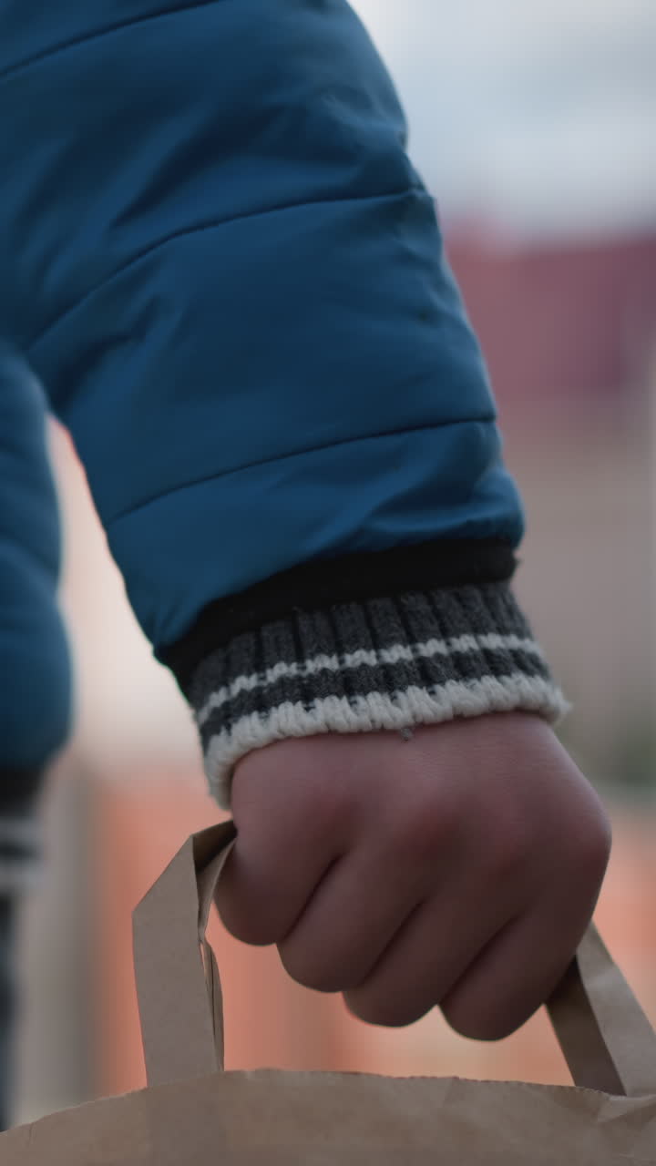Rear view of an individual in blue clothing holding a shopping bag, with a focus on the hand gripping the bag against a blurred suburban backdrop