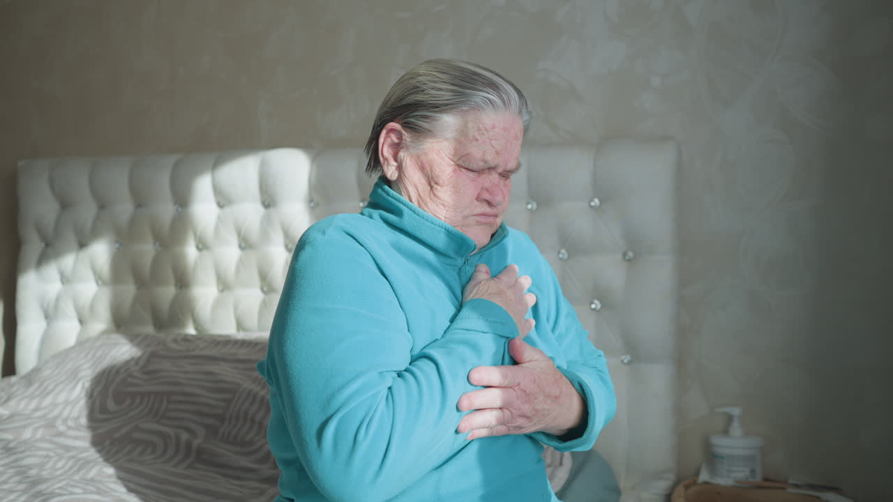 Elderly woman in green experiencing chest pain, gripping chest, eyes closed in discomfort, sitting on bed, sunlight coming through window, feeling unwell, distressed, physical struggle