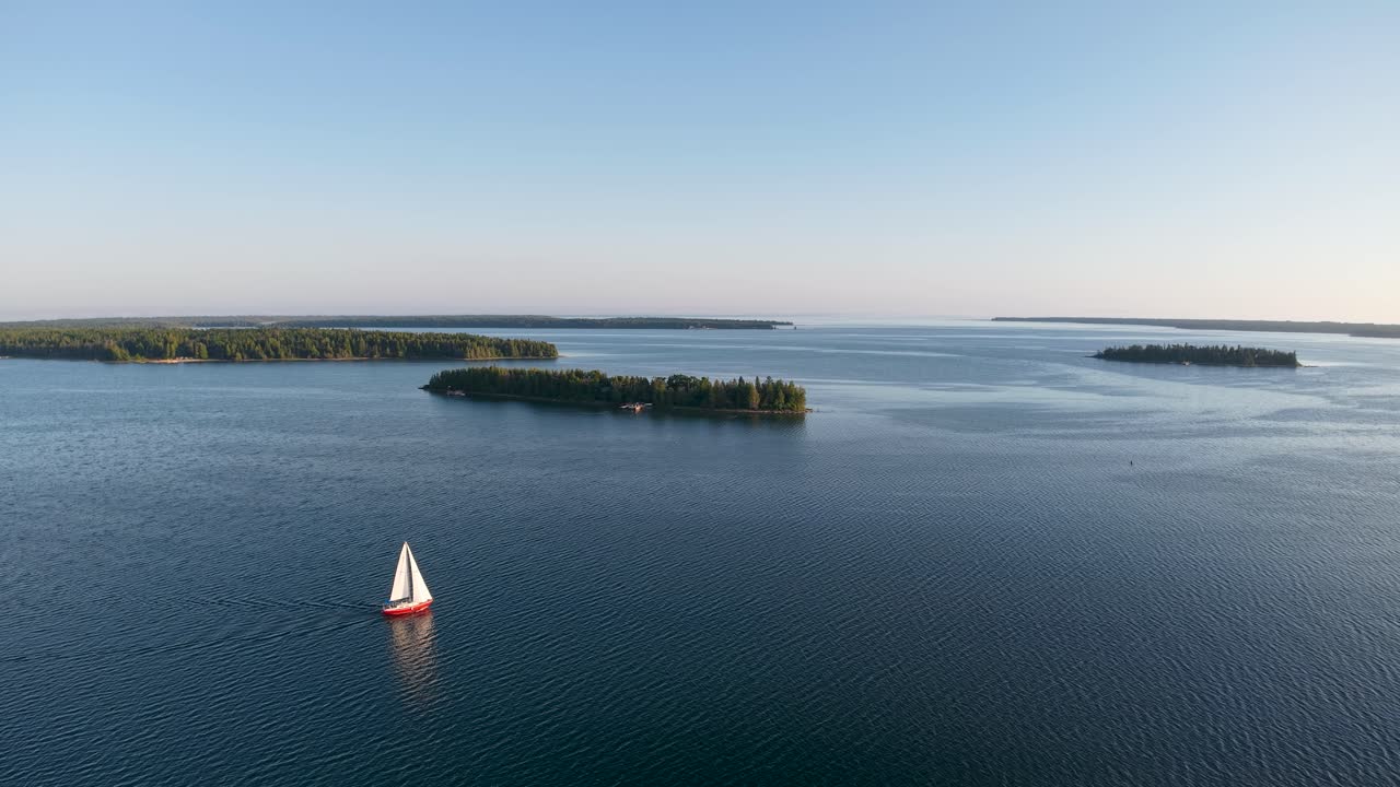 Aerial drone footage of a small sailboat gliding across the calm blue waters near forested islands in the Les Cheneaux Islands of Michigan’s Upper Peninsula