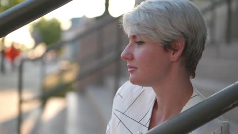 Strong-beautiful-women-in-fun-fashion-striped-white-suit-jacket-top-sitting-on-stairs-leaning-against-rail-looking-off-in-distance,-then-into-camera