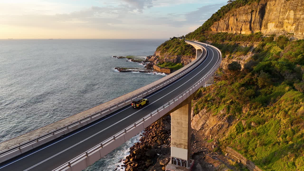 Spectacular aerial shot of a car on the Sea Cliff Bridge, highlighting the dramatic coastline and winding roadway. Tracking drone footage.
