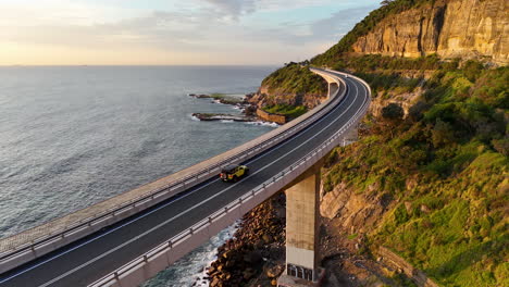 spectacular aerial shot of a car on the sea cliff bridge, highlighting the dramatic coastline and winding roadway. tracking drone footage.