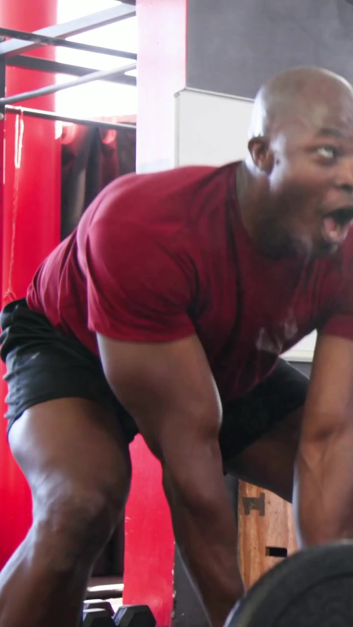 Vertical video: Bending man at gym in red tee black shorts grasping barbell and doing reps to train