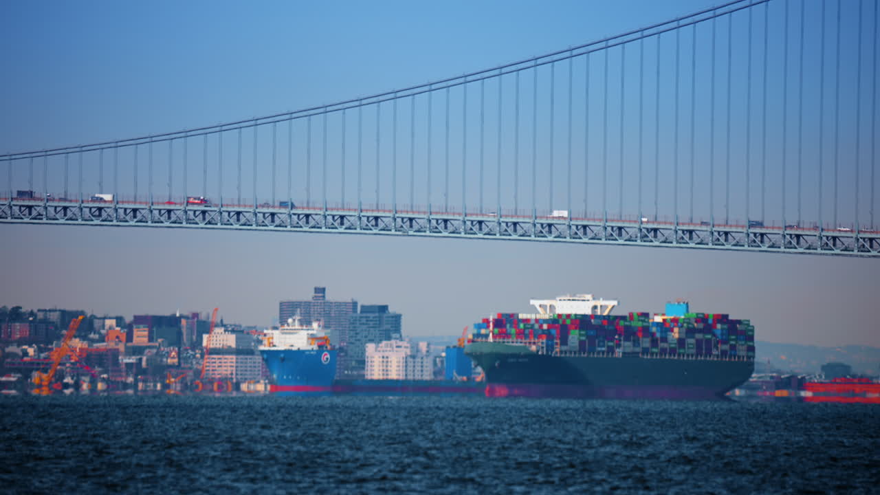 New York, USA, 25 December 2025: Large container ship sailing under suspension bridge in New York. Cargo ship transporting colorful containers on water under bridge structure