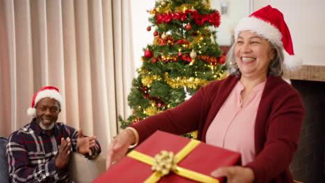 Feliz-Pareja-De-Ancianos-Diversos-Con-Sombreros-De-Navidad-Intercambiando-Regalos-Y-Abrazándose-En-Casa,-Cámara-Lenta