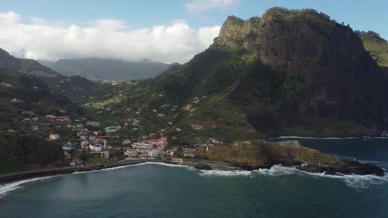 Riprese aeree mozzafiato catturano le lussureggianti scogliere dell'isola di Madeira che si ergono sopra un villaggio costiero con tetti rossi, con le onde che si infrangono contro le coste scoscese nella morbida e dorata luce del mattino