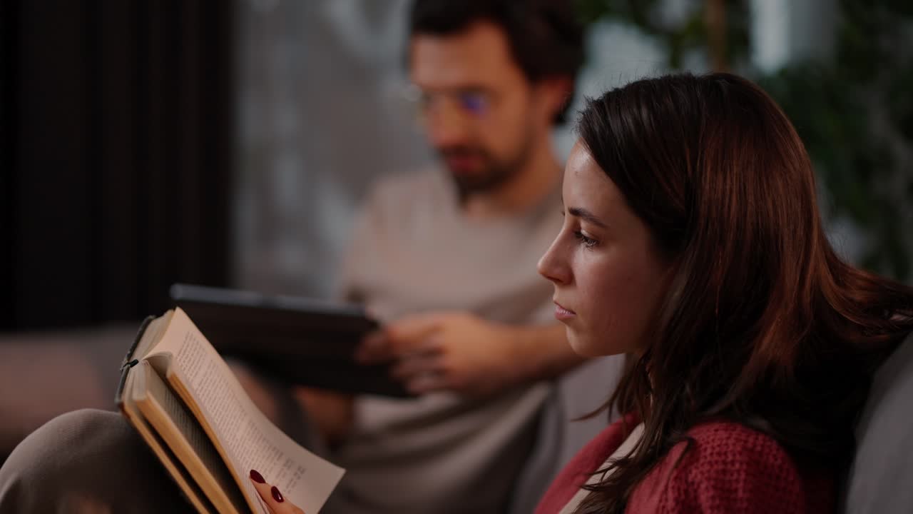 Side view of a confident brunette girl in a red sweater reading a book while sitting on the sofa with her boyfriend who is busy with a tablet in a modern apartment