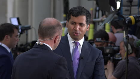 two television news anchors talk to each other as members of the press have gathered on downing street.
