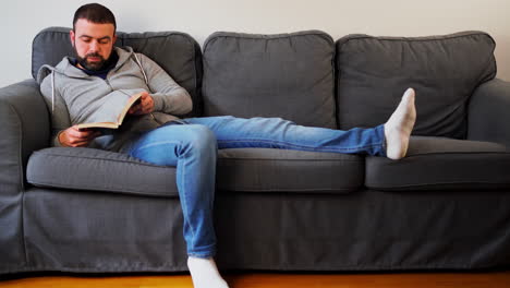 man reading book while sitting on the couch in his apartment