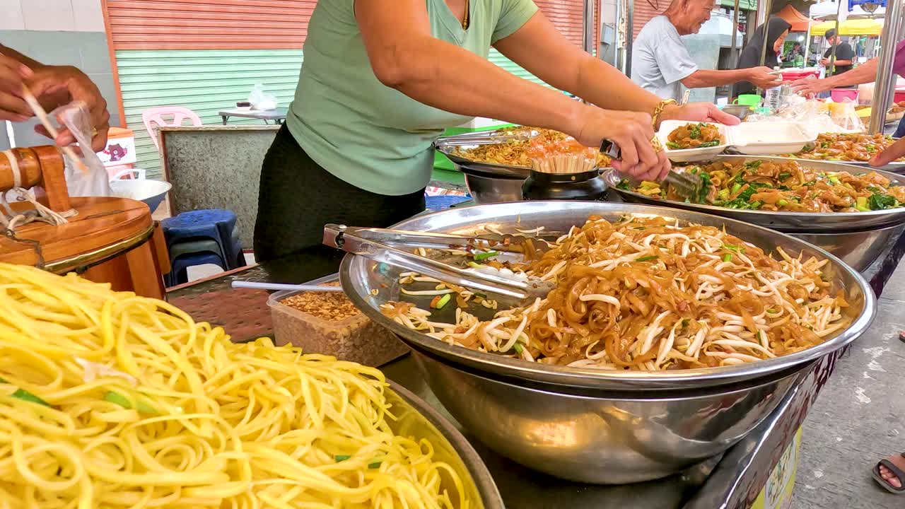 A bustling street vendor prepares vibrant noodle dishes in Kanchanaburi, Thailand. Bright lighting highlights the colorful ingredients and lively atmosphere