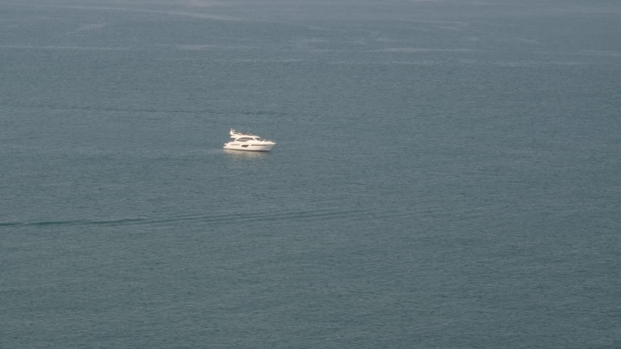 Locked-off wide shot of white yacht gently cruising on calm blue ocean surface