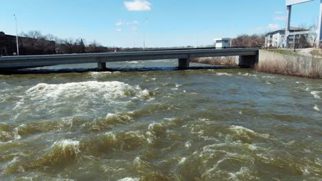 Flight-over-raging-Fox-River-going-underneath-a-bridge-in-Kaukauna,-Wisconsin