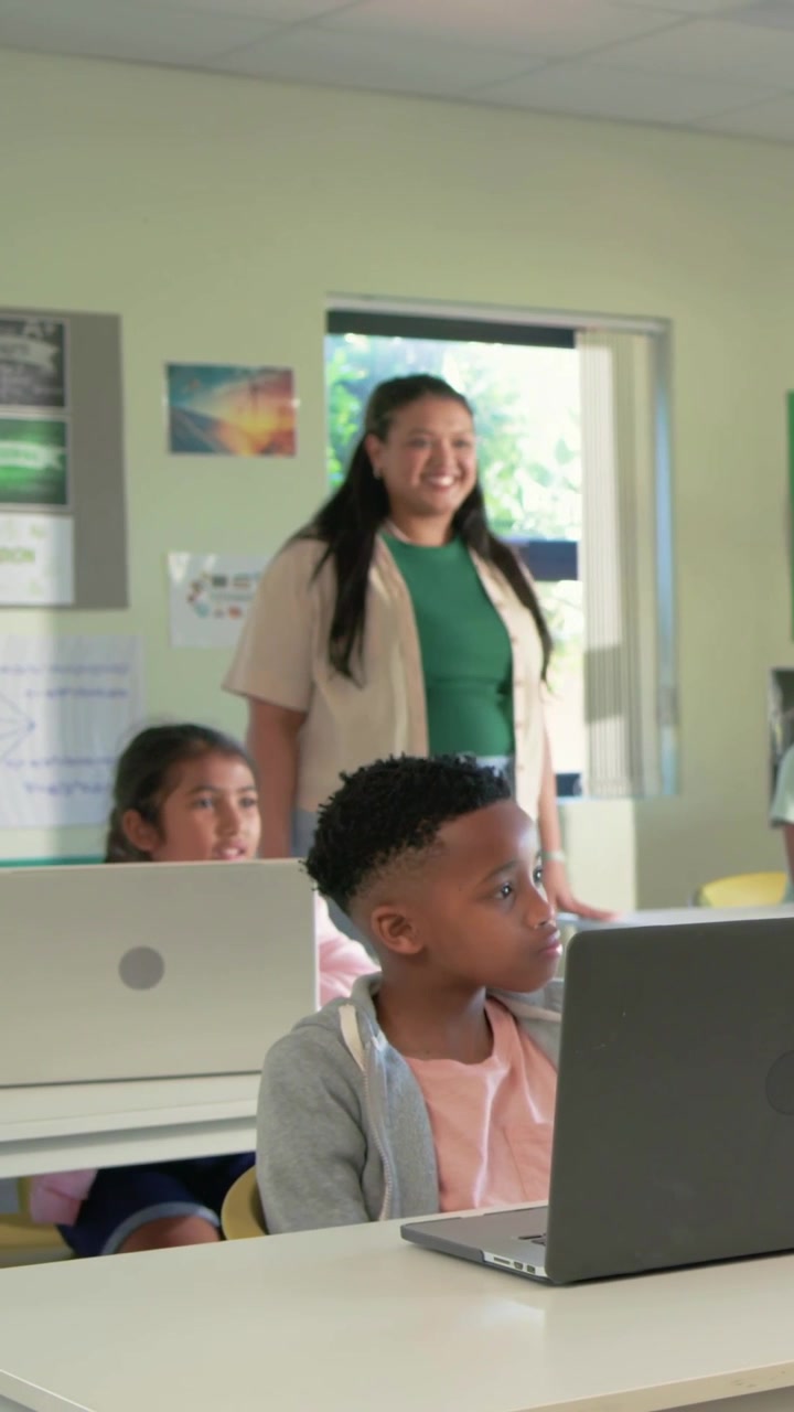 Video verticale: Insegnante in verde che entra e supervisiona un ragazzo che usa il laptop in classe, tablet, spazio per il testo.