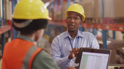 trabajadores afroamericanos masculinos y femeninos con casco y usando computadora portátil en el almacén