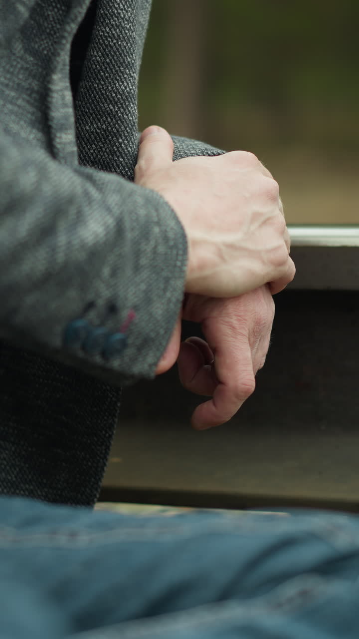 una vista de cerca de las manos de un hombre unidas, descansando en una vía ferroviaria. lleva un blazer gris y pantalones vaqueros azules