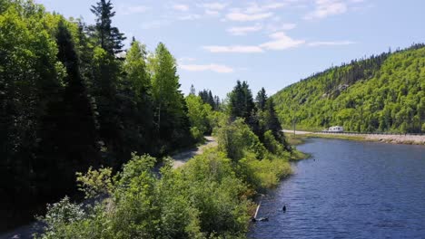 A-drone-flies-towards-a-highway-with-vehicles-over-a-lake-surrounded-in-a-forest-during-the-day-in-Ontario,-Canada