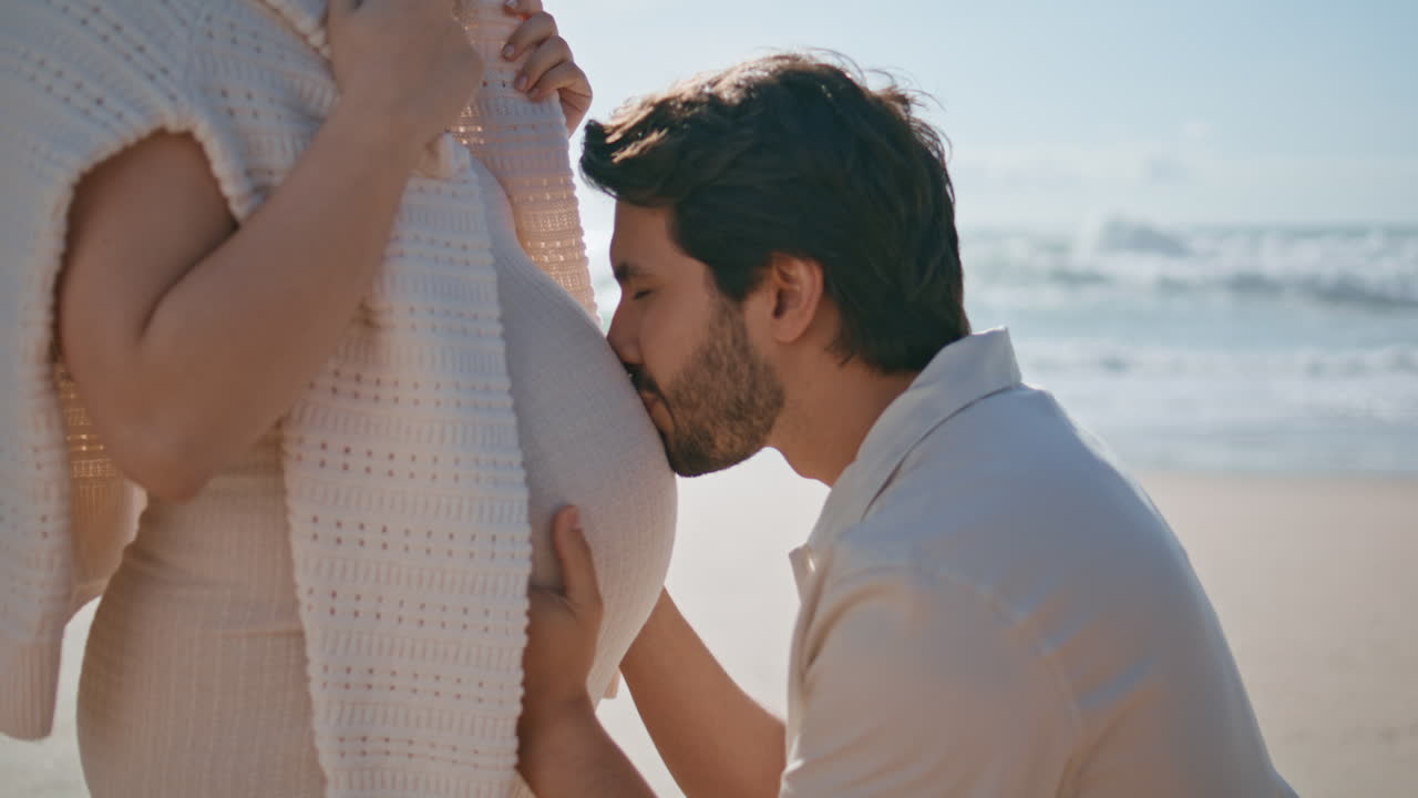 el marido besando a la esposa embarazada en el vientre de la playa soleada de cerca. el hombre expresando amor