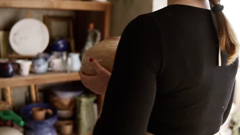 Side-View-Portrait-Of-Contemporary-Female-Potter-Putting-Handmade-Vase-On-Shelf-In-Studio,-Pleased-With-Her-Work,-Smiling-To-The-Camera