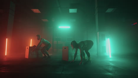 un hombre y una mujer saltan sobre una caja realizando un burpee. trabajo en equipo conjunto. pareja atlética realiza ejercicio cardiovascular salto funcional burpee en el gimnasio en colores oscuros de neón