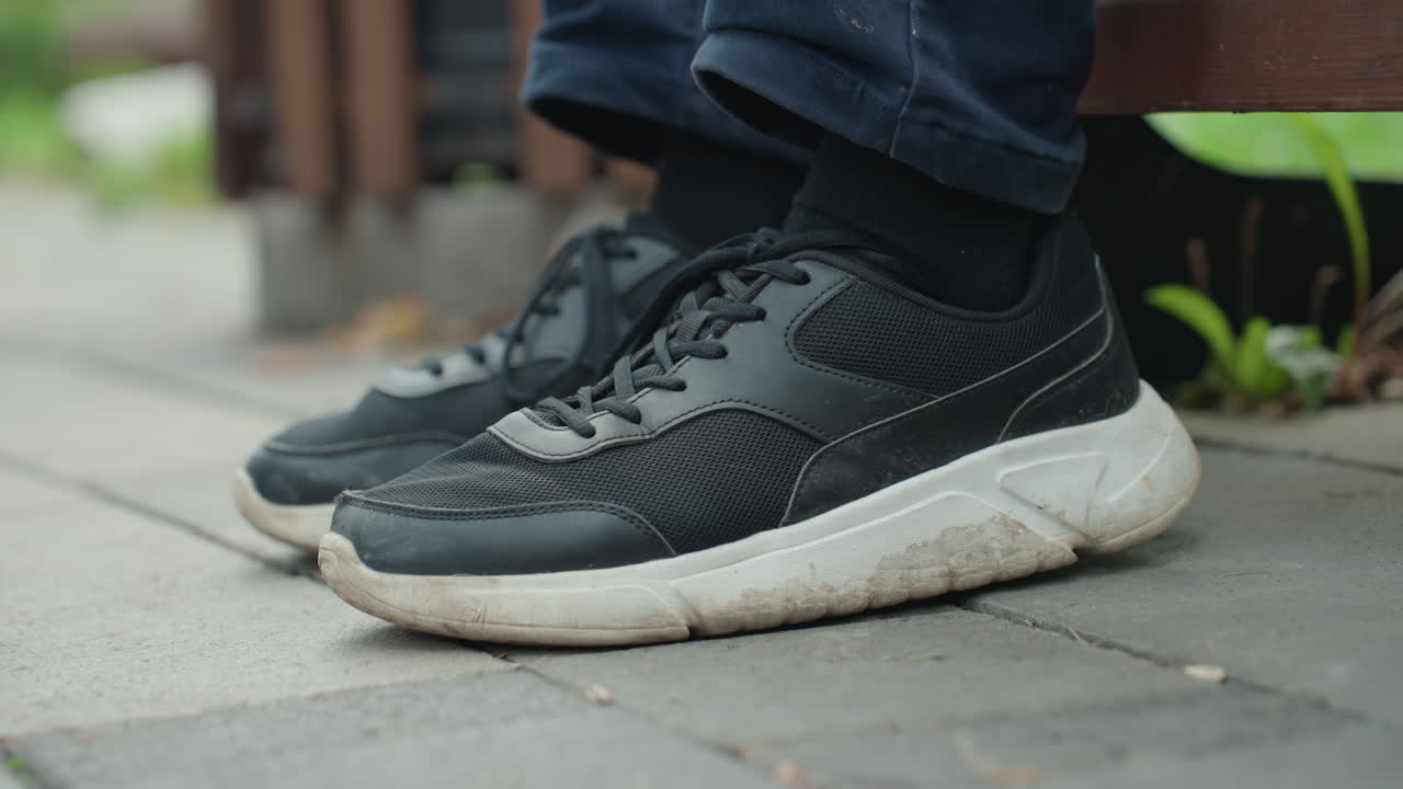Nærbillede af sort sneaker på fortov viser afslappet pendler, der hviler fødderne ved siden af en bænk, slidt hvid sål, strikoverdel, snøredetalje, denimkant og sok synlig, lav dybdeskarphed, dæmpede byfarver