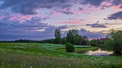 Timelapse-shot-of-clouds-passing-by-in-timelapse-over-wooden-cottage-by-the-side-of-a-lake-in-distance-during-evening-time