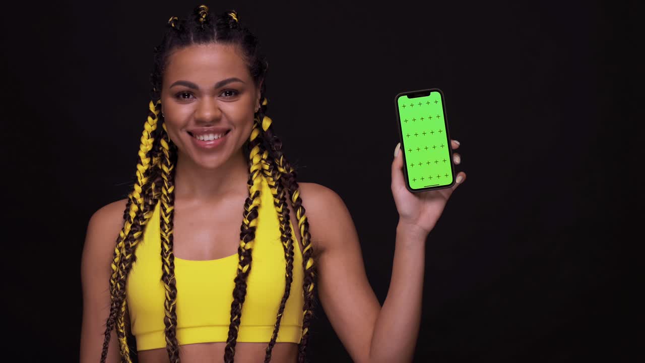 retrato de una chica afroamericana sonriente apuntando con el dedo a la pantalla del teléfono móvil con una llave cromática. mujer aislada sobre un fondo negro