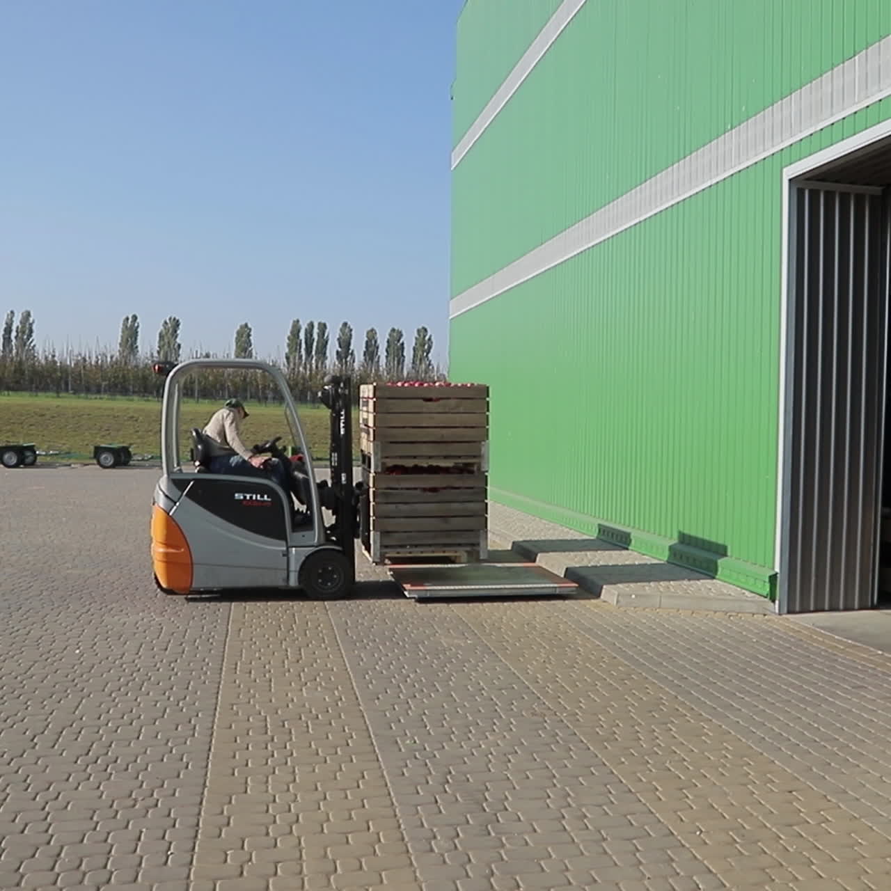 A worker delivers the two wooden boxes with ripe apples to a pallet in the street near the entrance to the warehouse of the juice processing plant.