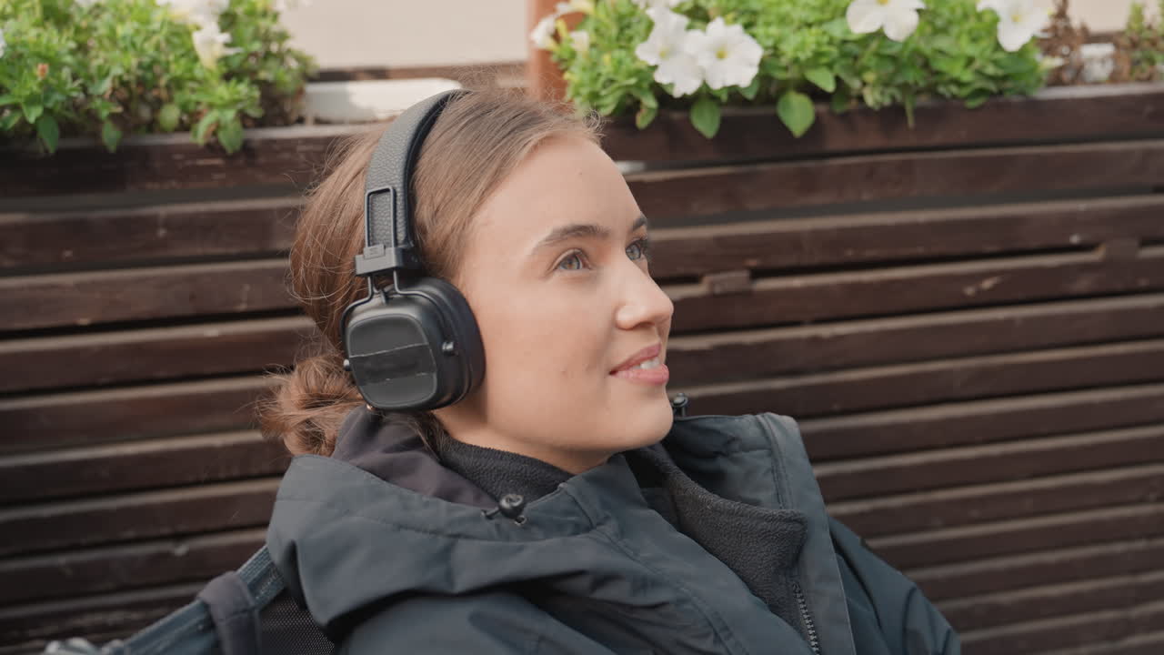 Giovane donna su un balcone che ascolta musica, con una giacca comoda e cuffie wireless, appoggiata all'angolo di una fioriera con fiori bianchi, luce soffusa, espressione serena, scene che suggeriscono uno stato d'animo riflessivo, ideale per uno studente